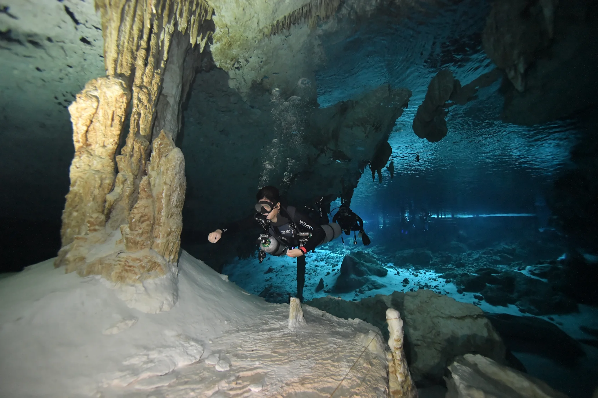 Buceador en caverna subacuática iluminada por haces de luz natural con formaciones rocosas y visibilidad cristalina<br />
