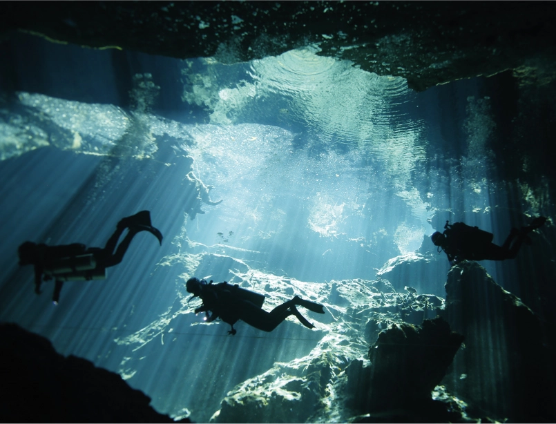 Buceador en caverna subacuática iluminada por haces de luz natural con formaciones rocosas y visibilidad cristalina<br />
