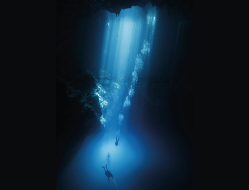 Buceador en caverna subacuática iluminada por haces de luz natural con formaciones rocosas y visibilidad cristalina<br />
